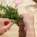 先以鐵絲穿過藤圈並收好，抓一把雪松，繞過後拉緊，再取一把雪松，在第一把根部接上，以此將雪松綁完。